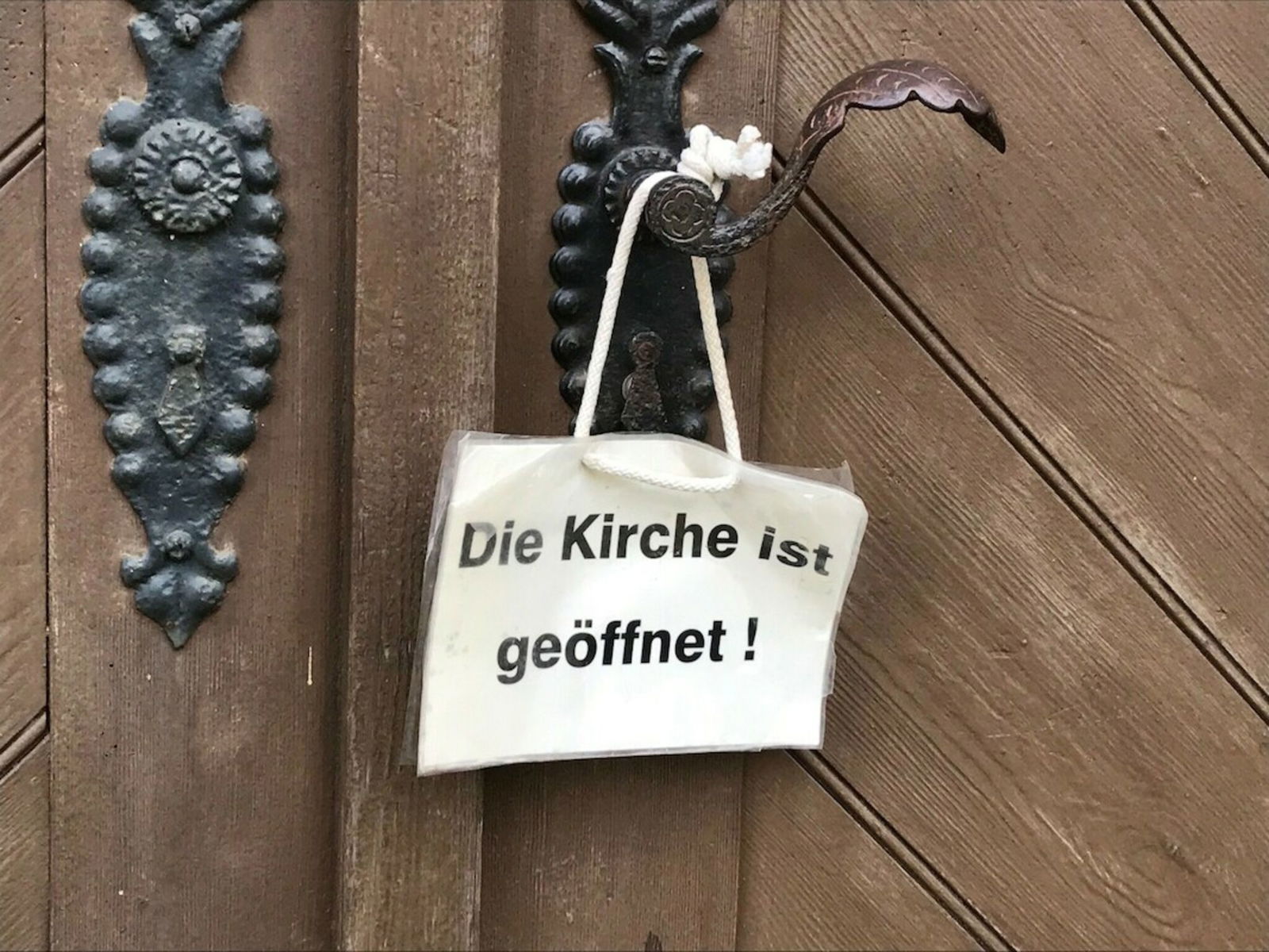 An einer Kirchentür hängt ein Schild. Darauf steht: Die Kirche ist geöffnet