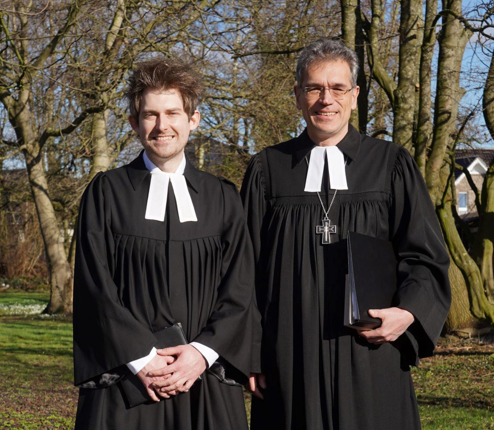 Zwei Pastoren im Talar stehen in einem Park. Sie lächeln freundlich in die Kamera.