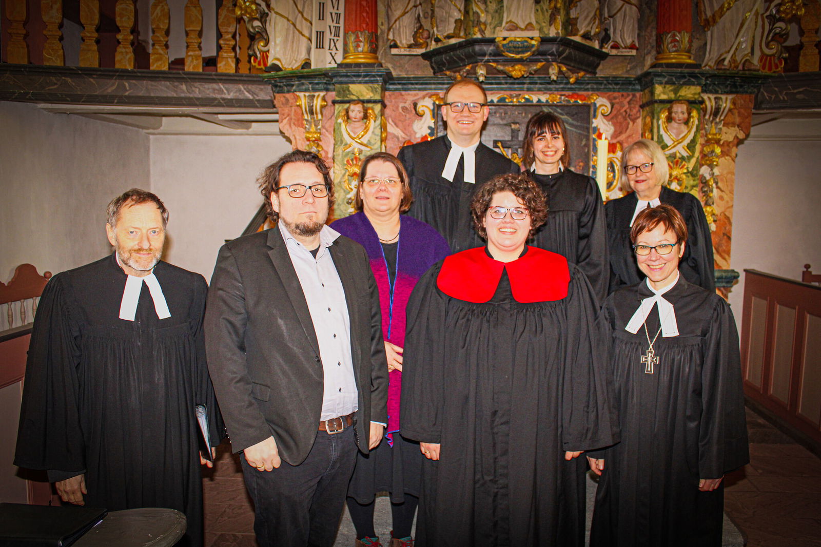 Eine Gruppe von Menschen steht vor einem Alter. Einige tragen Talare. Sie lächeln freundlich in die Kamera.