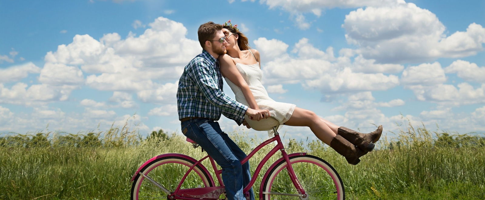 Das Bild zeigt zwei Personen auf einem Fahrrad, eine davon sitzt vorne auf dem Lenker. Das Paar fährt durch die Natur, im Hintergrund ist ein grünes Feld zu sehen. Am blauen Himmel sind einige weiße Wolken.