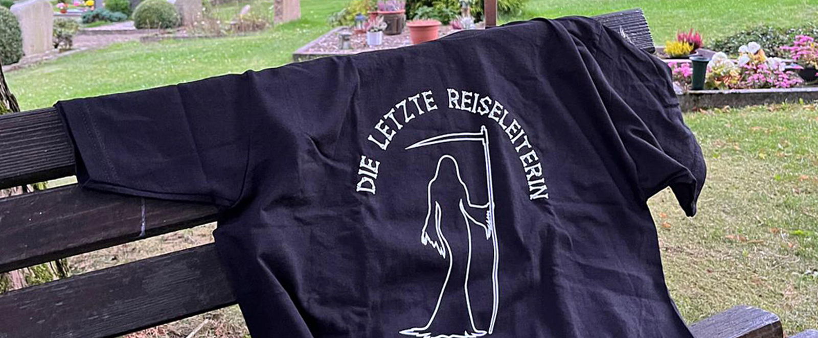Ein schwarzes T-Shirt mit der Aufschrift „Die letzte Reiseleiterin“ auf einer Friedhofsbank. 