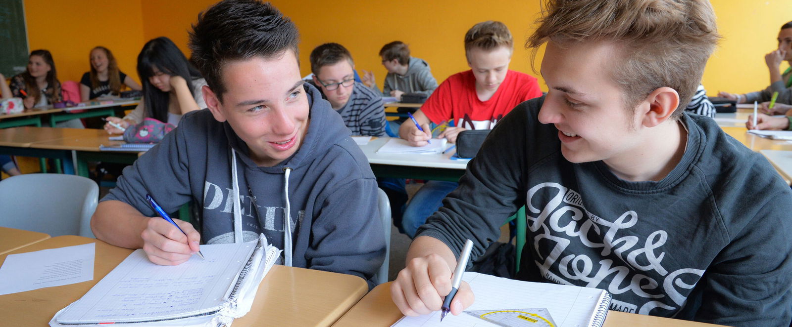 Zwei Schüler sitzen in einem Klassenraum vor ihren Heften und lächeln sich an.