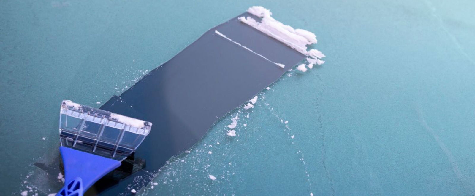 Ein Eiskratzer mit blauem Griff liegt auf einer vereisten Autoscheibe, auf der eine kleine Bahn freigeschoben wurde.