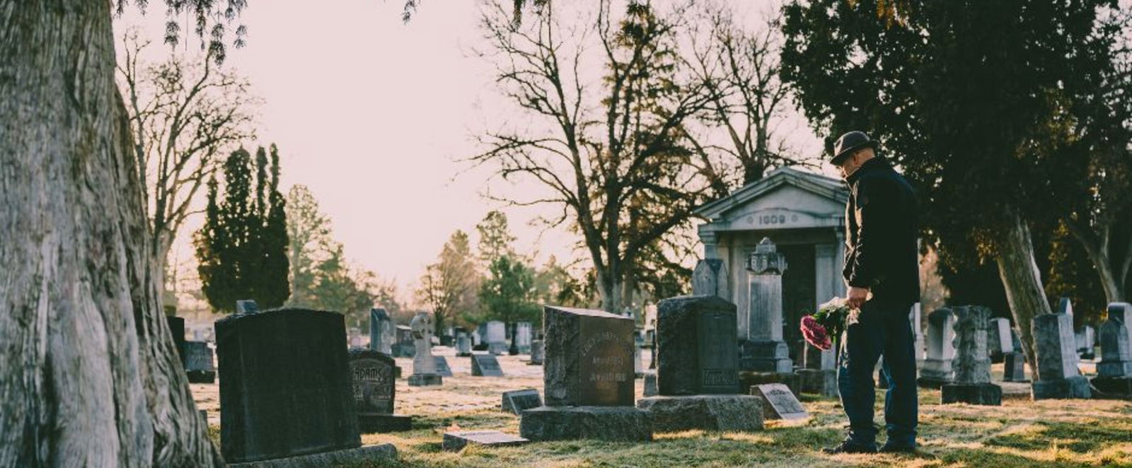Ein Mann geht auf einem Friedhof zu einem Grab. Er hält einen Straß mit Blumen in der Hand. Sein Blick ist gesenkt.