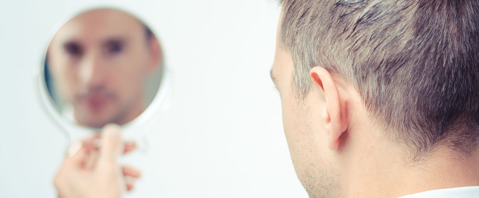 Ein Mann betrachtet sich und seine grauen Haare kritisch in einem Handspiegel. 