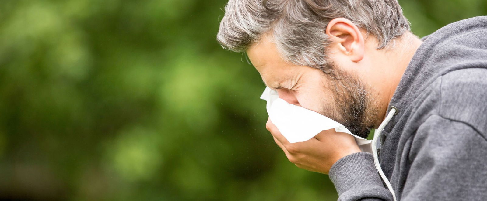Eine als Mann lesbare Person steht draußen im Grünen und schneuzt in ein Taschentuch.