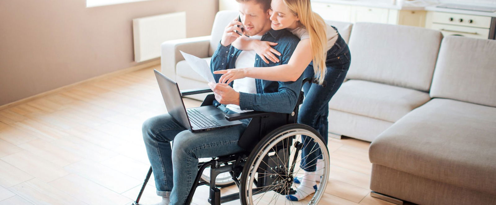 Eine als Frau lesbare Person umarmt von hinten eine als Mann lesbare Person im Rollstuhl, die am Laptop arbeitet.