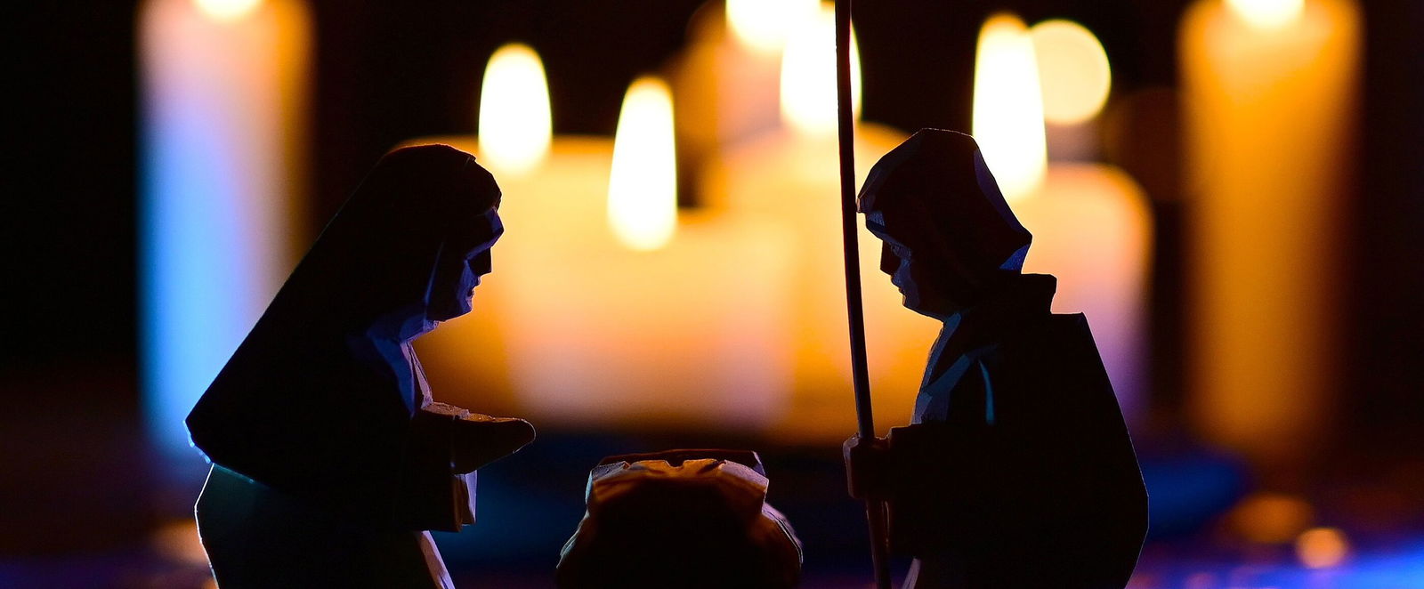 Die Umrisse einer Krippe und zweier kniender Figuren vor unscharfen brennenden Kerzen im Hintergrund