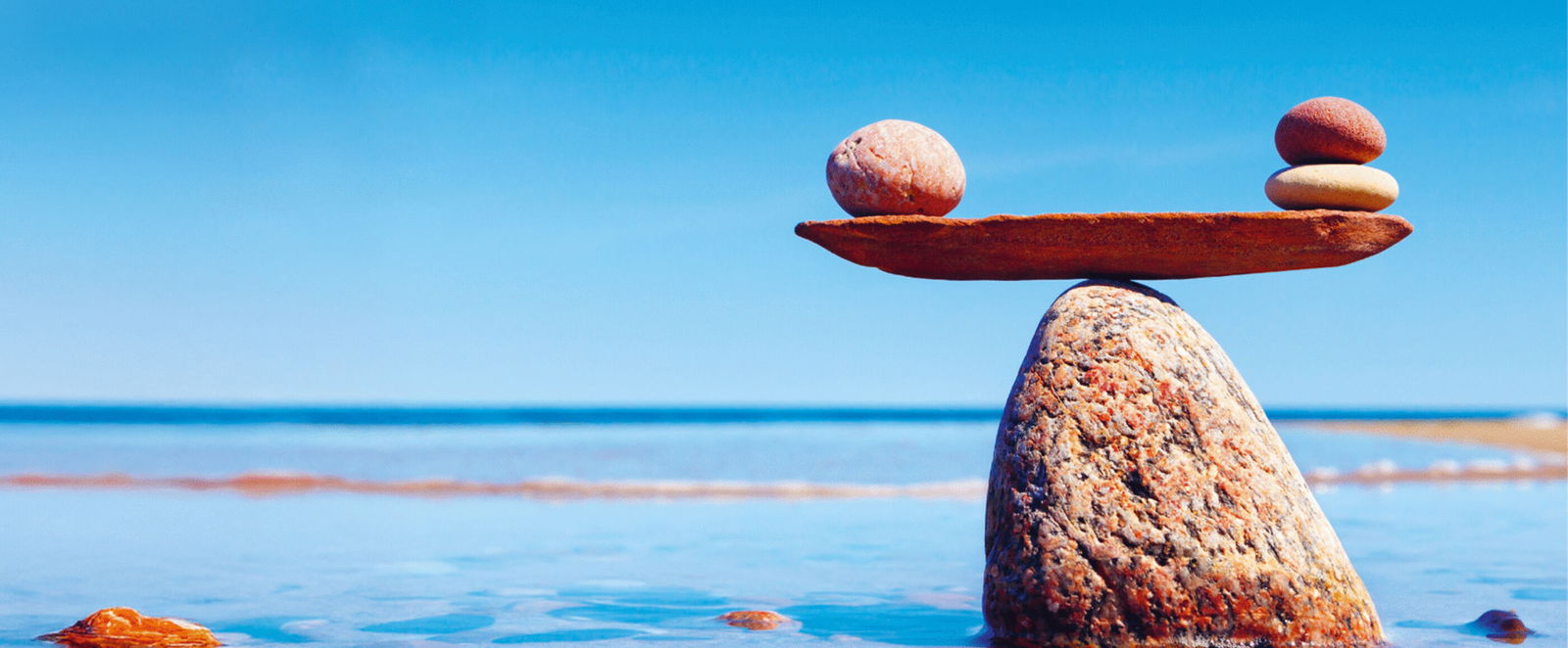 Ein großer Stein in flachem Wasser mit einem Brett darauf, auf dem kleinere Steine in einer Waage balancieren. Daneben steht: Soviel du brauchst.