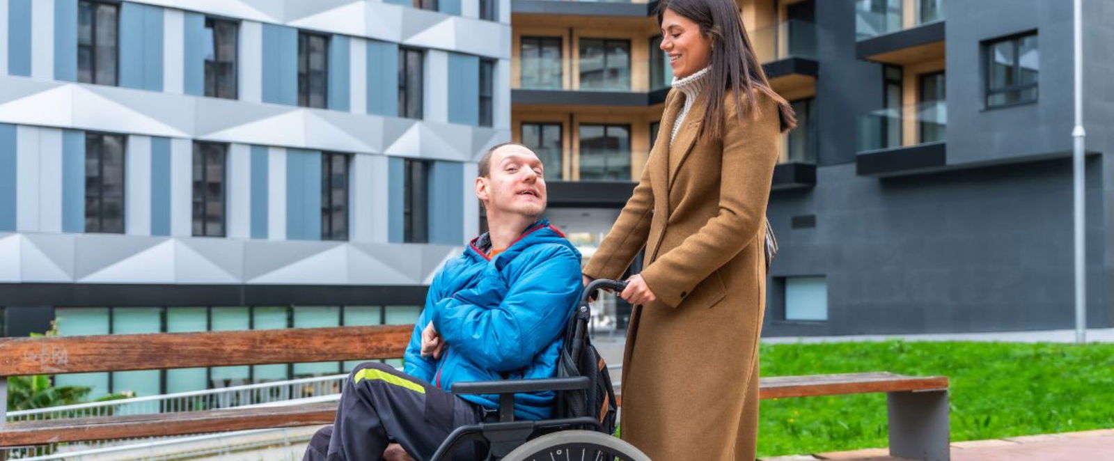 Eine junge als Frau lesbare Person schiebt einen jungen als Mann lesbare Person im Rollstuhl.