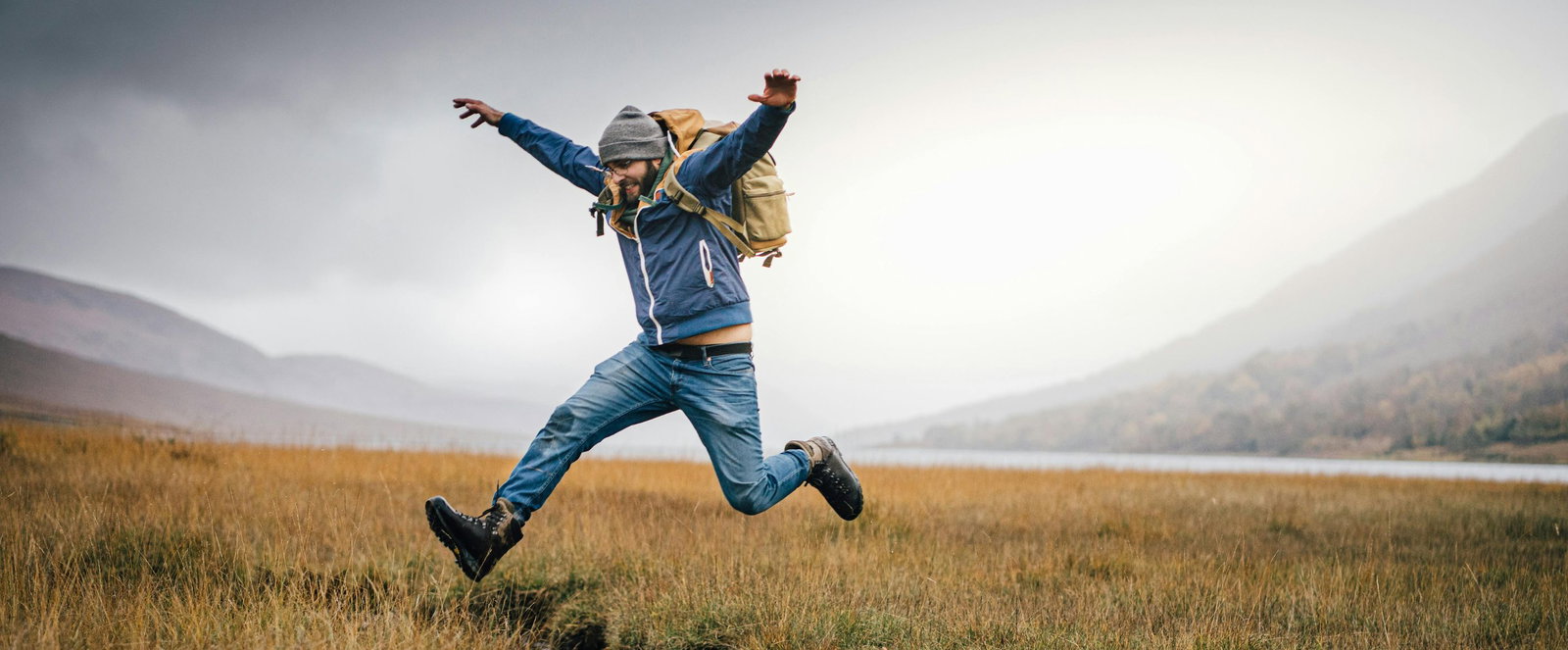 Ein Mann in Wandersachen und Rucksack springt auf einem Feld über einen Bachlauf.