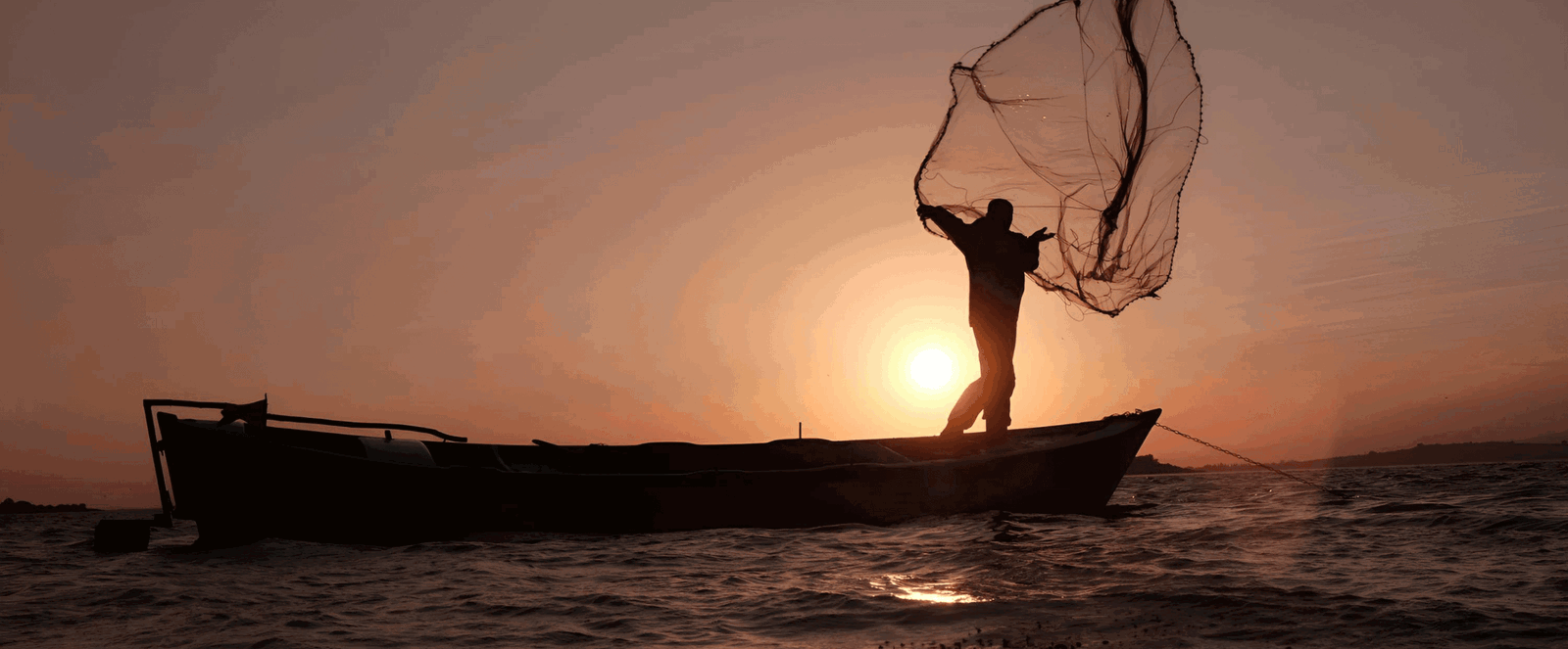 Ein Fischer wirft ein Netz von einem kleinen Boot aufs Meer vor einem Sonnenuntergang.