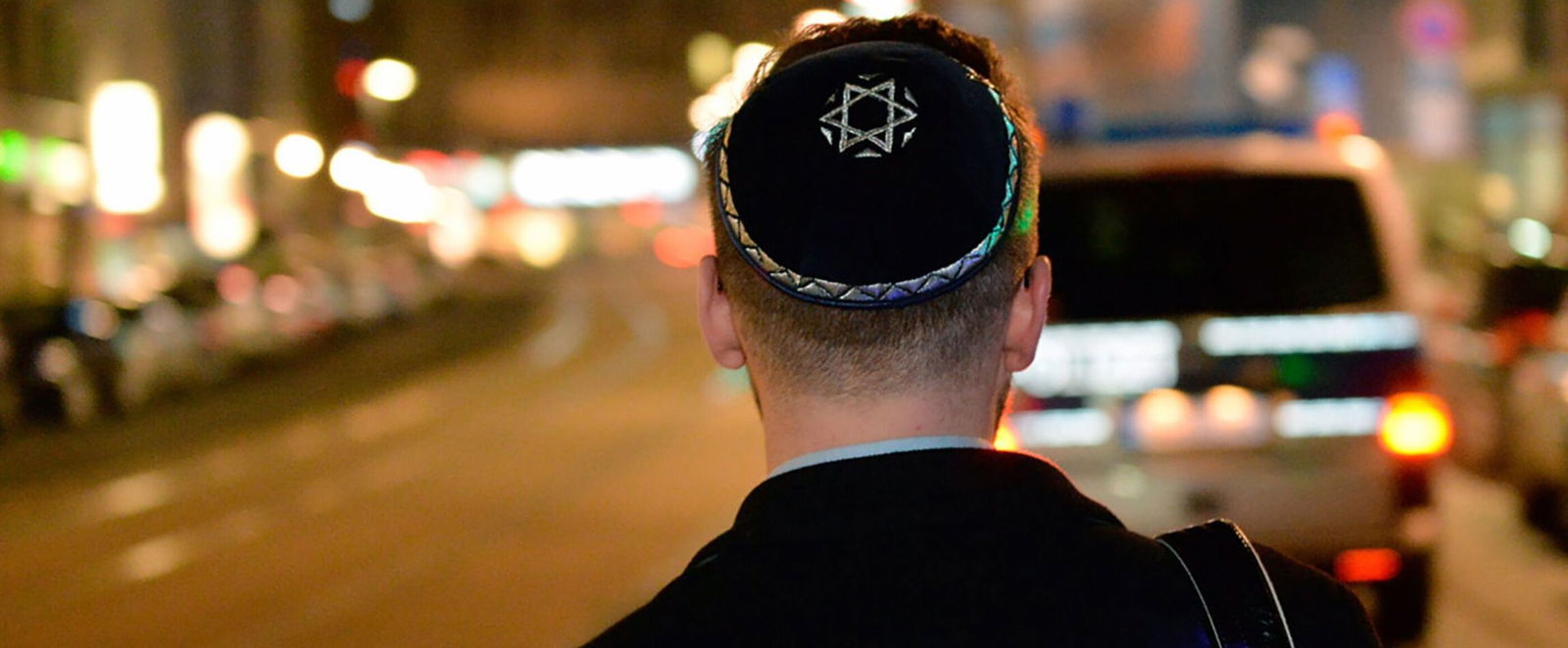 Ein Mann mit Kippa geht am Abend durch eine Stadt. Im Hintergrund sieht man ein Auto und Leuchtreklamen.