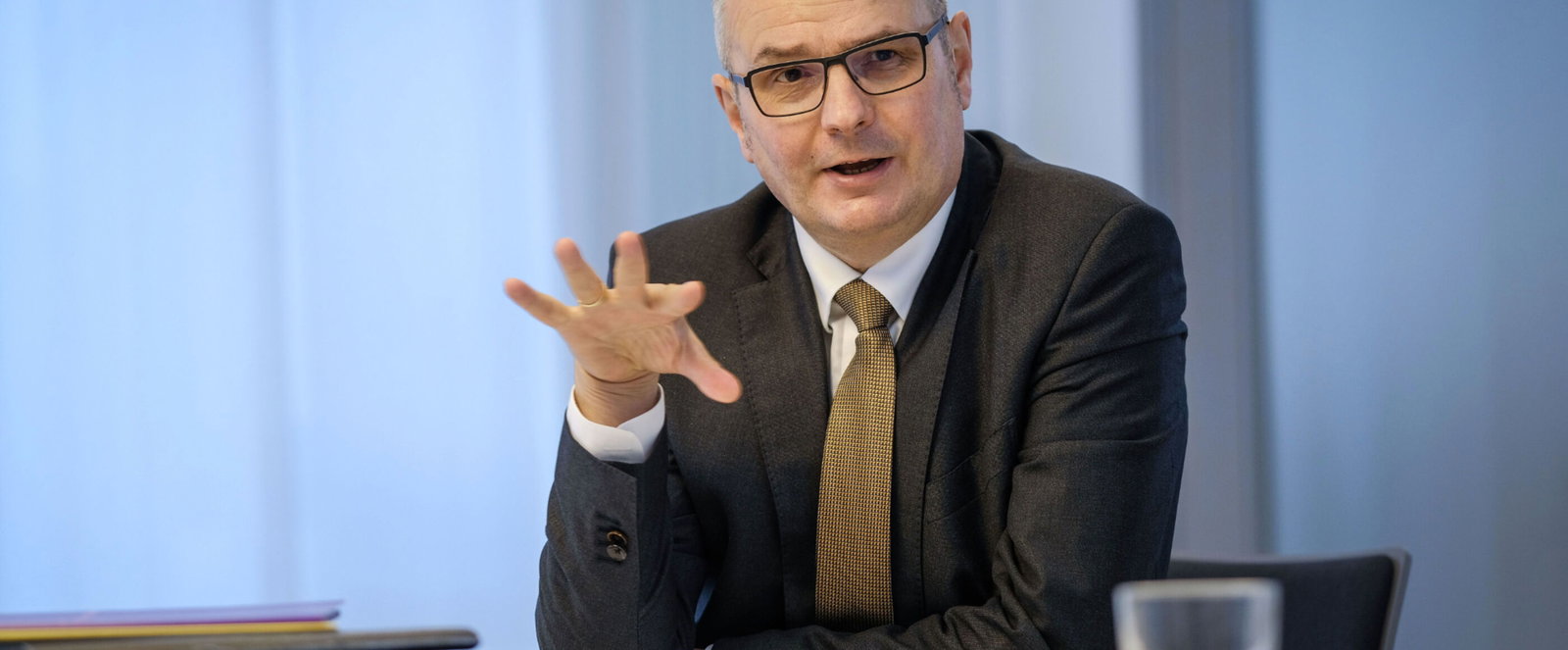 Ein Mann in einem dunklen Anzug sitzt an einem Tisch und erklaert etwas. Vor ihm liegen Akten, daneben steht ein Glas Wasser. Das Bild entstand bei einer Pressekonferenz.
