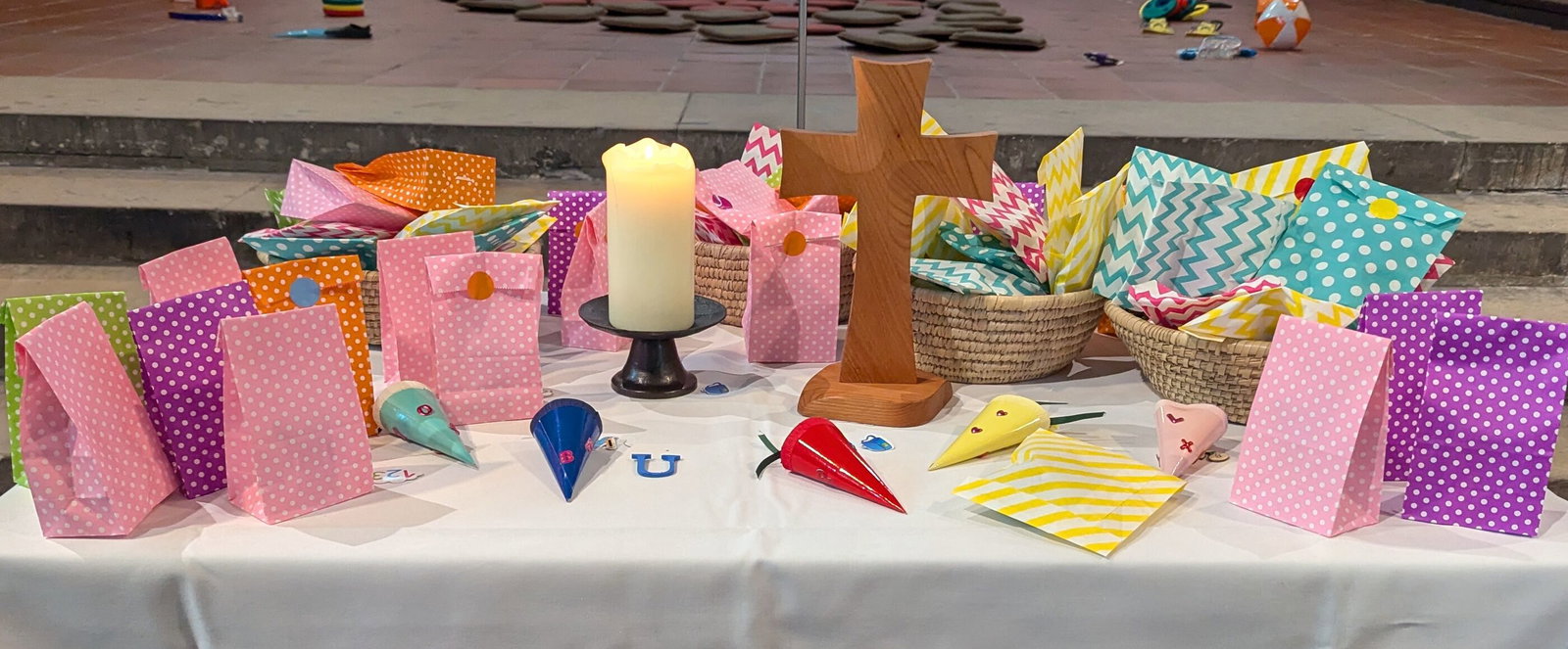 Auf einem Tisch stehen ein Kreuz aus Holz und mehrere bunte Papiertüten.