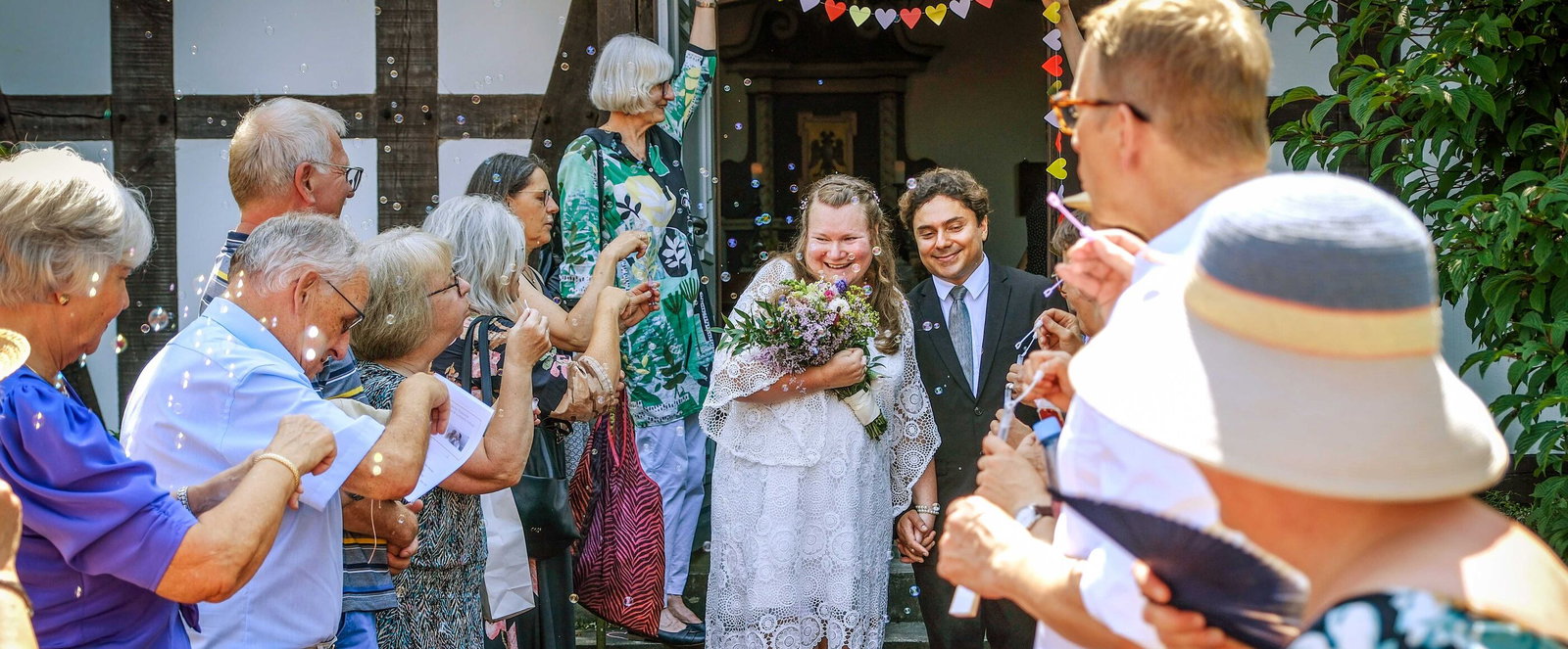 Vor einem Fachwerkhaus geht ein Hochzeitspaar durch eine Reihe freudiger Menschen, die Seifenblasen pusten.