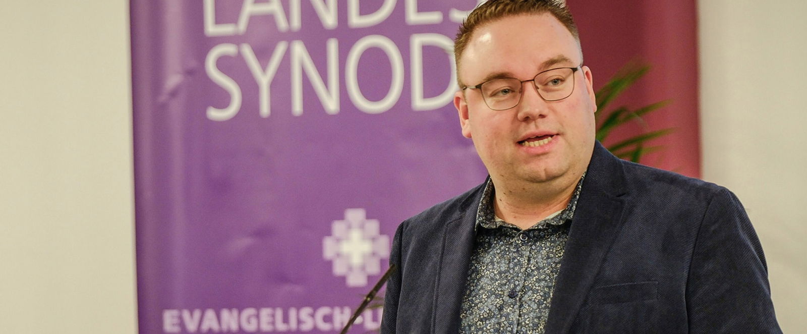 Eine männlich lesbare Person steht an einem Rednerpult vor einem Plenum