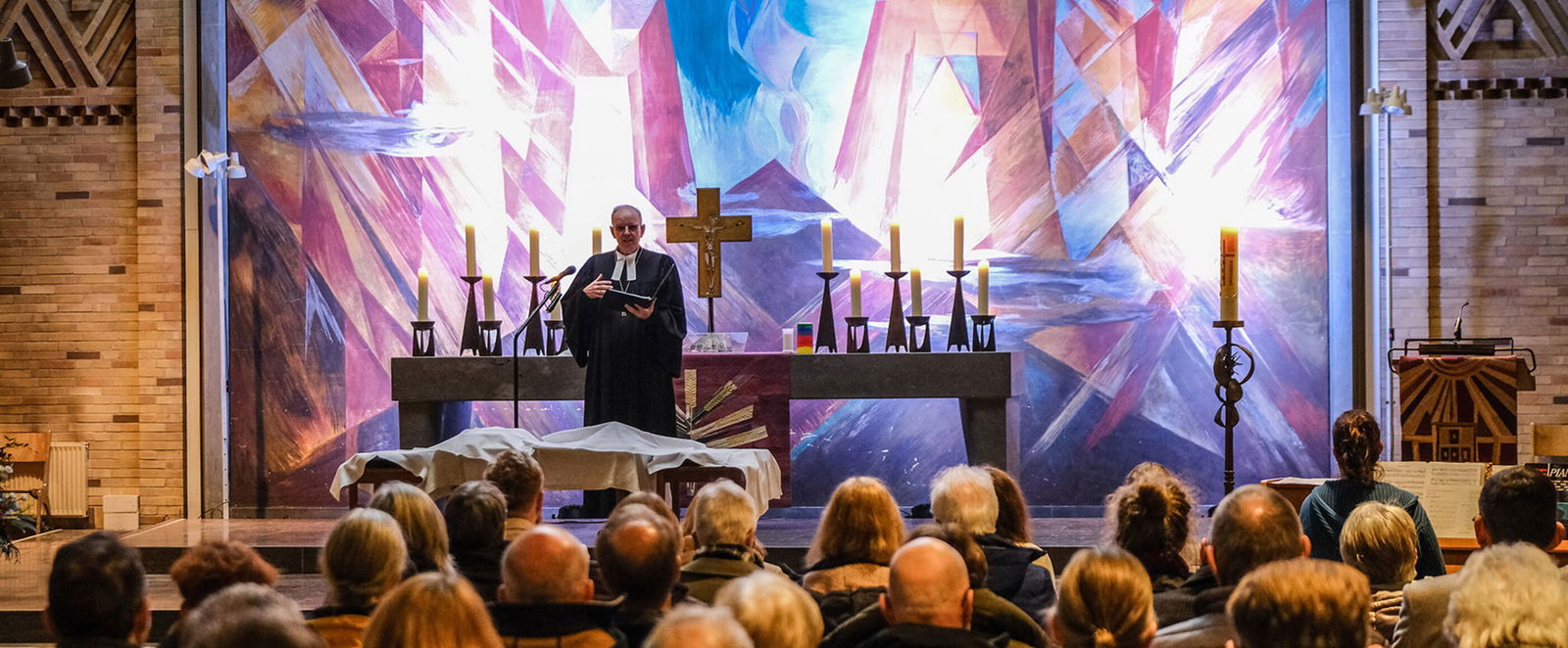 Männlich und weiblich zu lesende Personen sitzen in einem Kirchenraum und schauen auf eine männlich zu lesende Person im Talar, die im Altarraum steht und etwas vorliest.