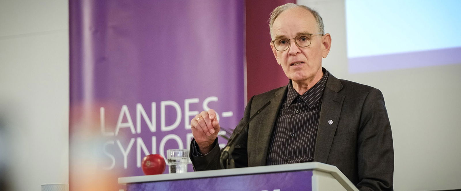 Eine männlich gelesene Person mit kurzen grauen Haaren und Brille hinter dem Redepult