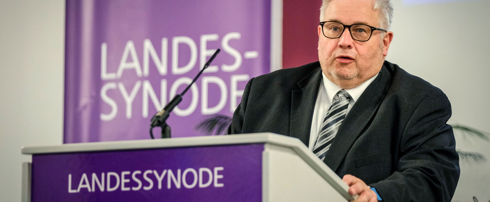 Eine männlich lesbare Person im Anzug steht am Rednerpult und spricht.