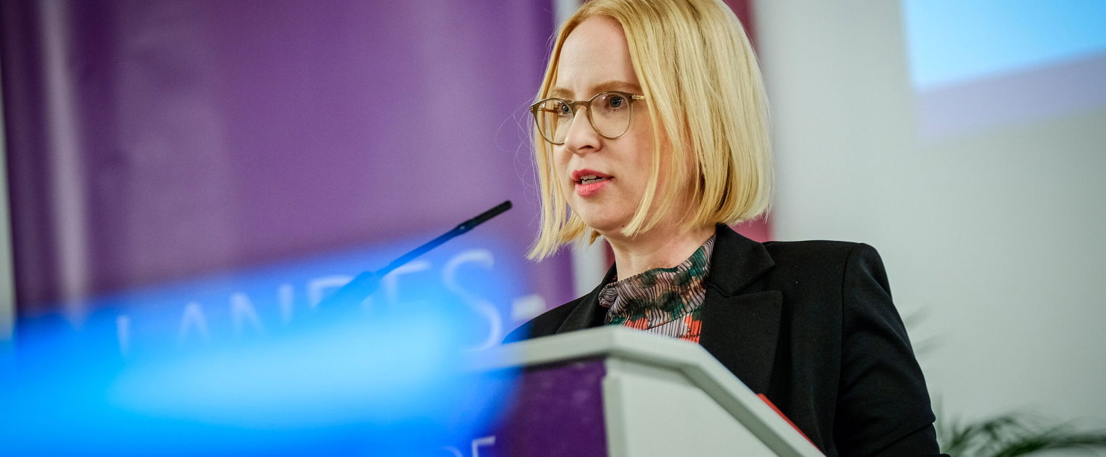 Eine weiblich gelesene Person mit blonden Haaren und Brille am Podium. 