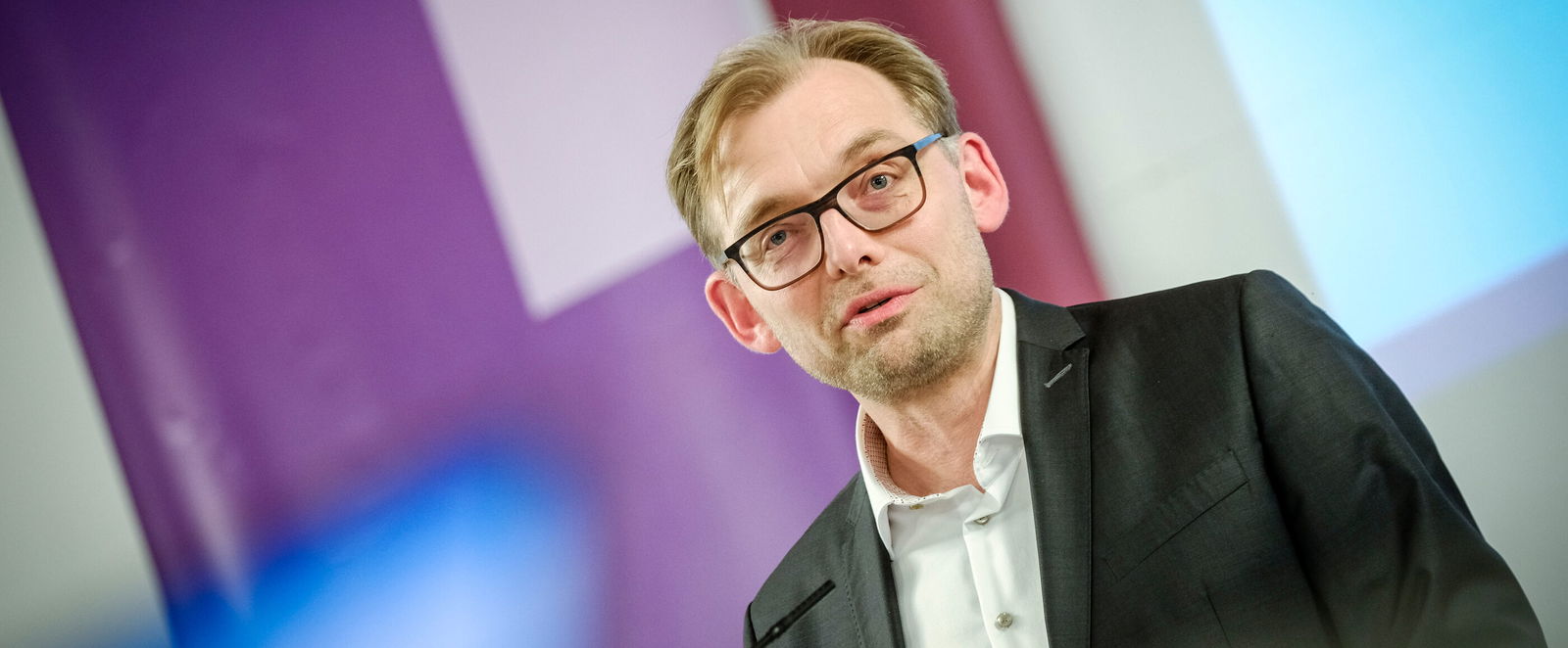 Eine männlich lesbare Person im Anzug mit Brille steht am Podium und spricht.