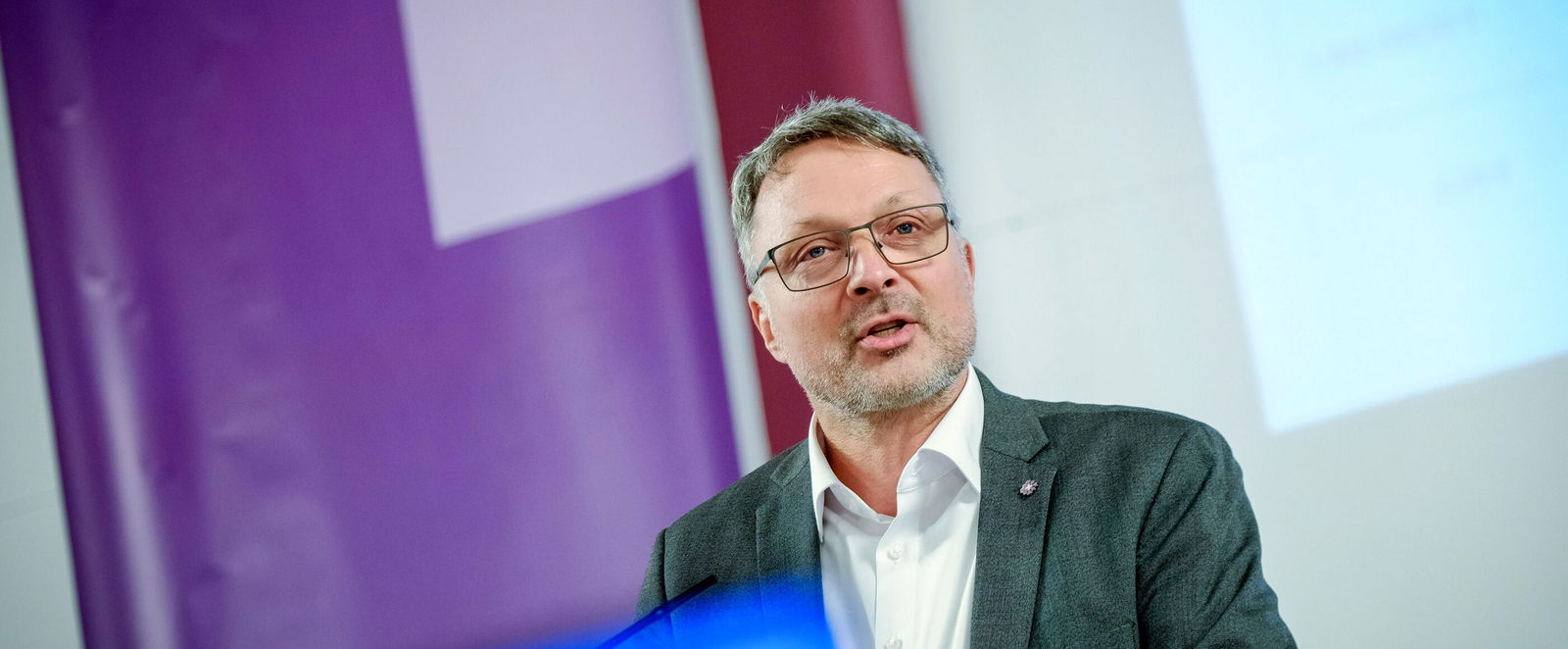 Eine männlich gelesene Person mit Brille und kurzen grauen Haaren am Podium.