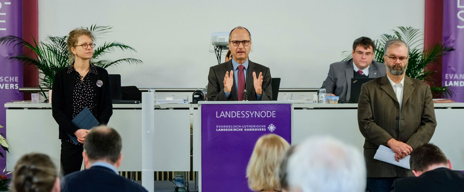 Eine männlich lesbare Person an einem Rednerpult und zwei Personen links und rechts daneben