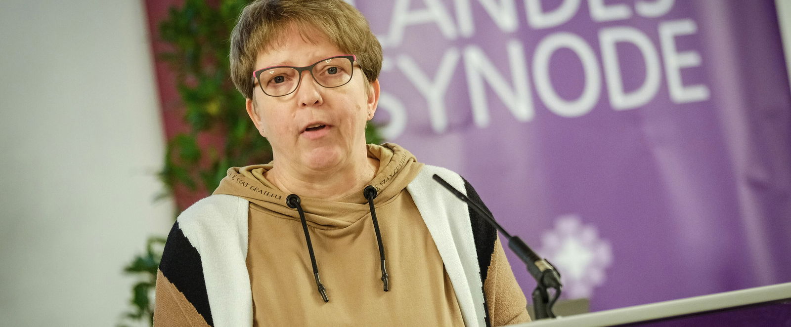 Eine weiblich lesbare Person mit Brille und kurzen Haaren steht hinter einem Rednerpult und spricht.