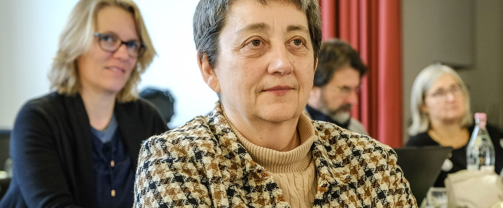 Eine weiblich lesbare Person mit karierter Bluse sitzt, mehrere weiblich lesbare Personen im Hintergrund