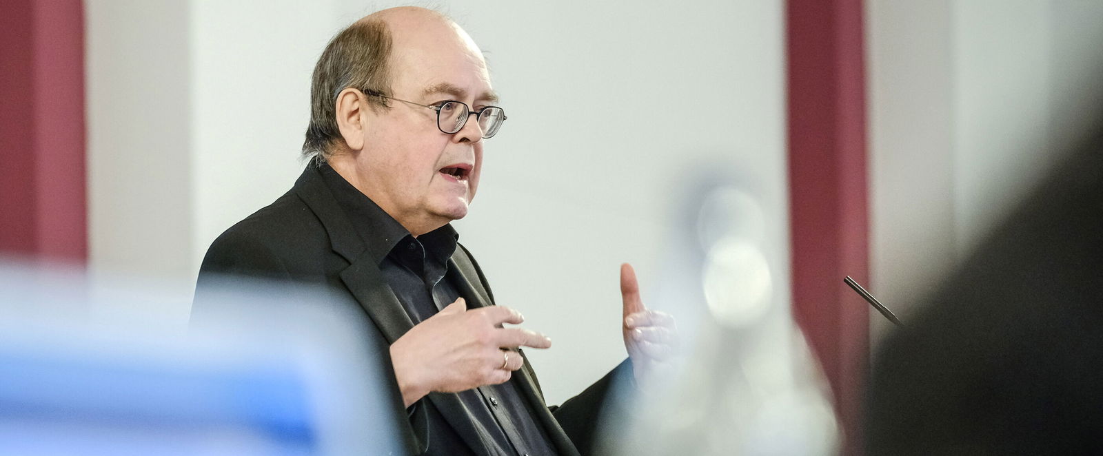 Eine männlich lesbare Person  mit Brille steht und gestikuliert 