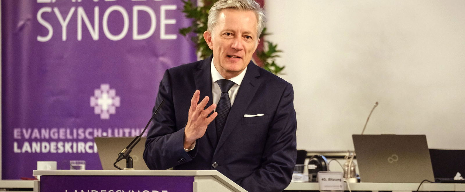 Eine männlich lesbare Person steht an einem Rednerpult und hebt eine Hand und spricht lächelnd zu den Zuhörerinnen.