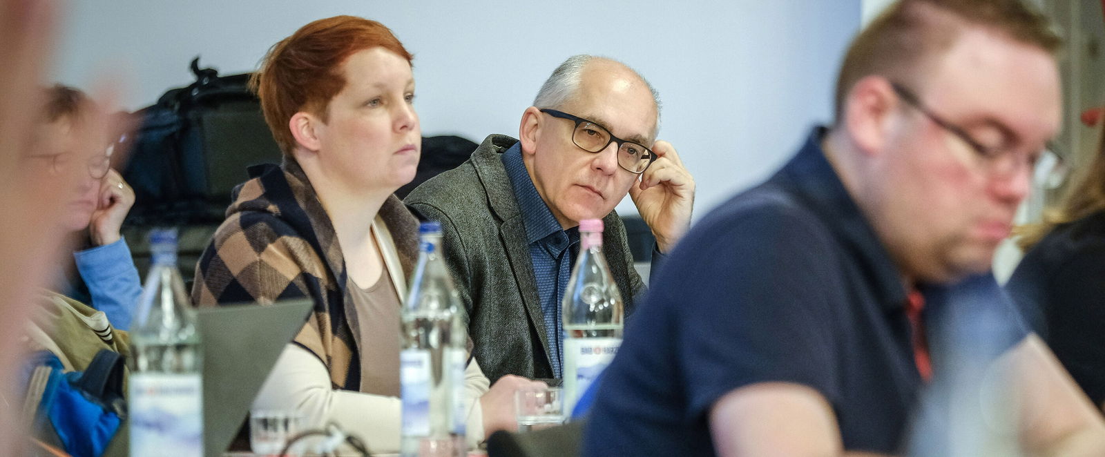 Eine männlich lesbare Person sitzt und schreibt, im Hintergrund weitere Personen, die zuhören