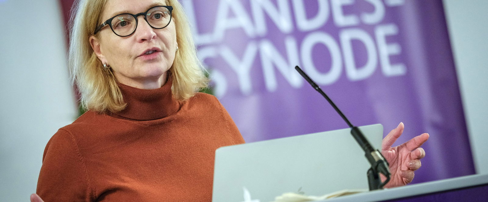 Eine weiblich lesbare Person mit Brille und einem Rollkragenpulliver steht an einem Pult und spricht.