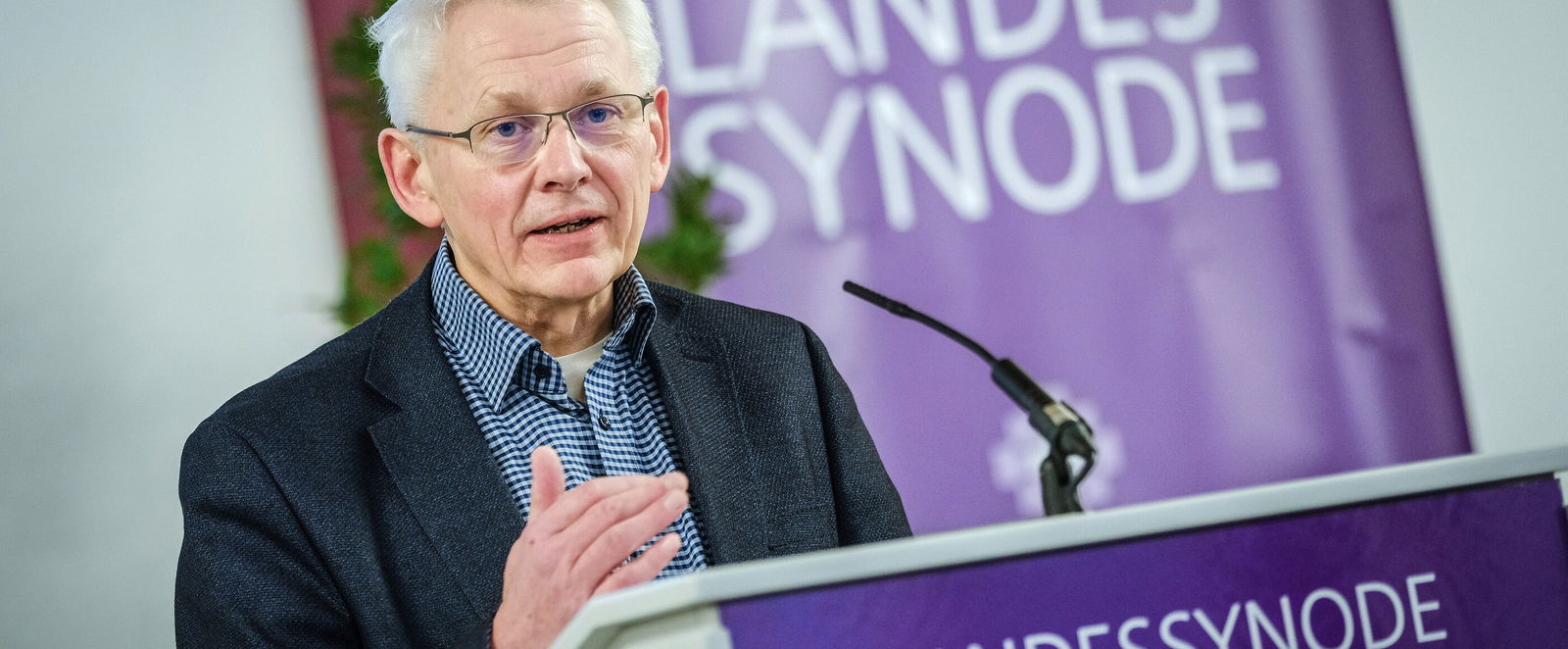 Eine männlich lesbare Person mit Brille und weissem Haar steht an einem Pult uns spricht.