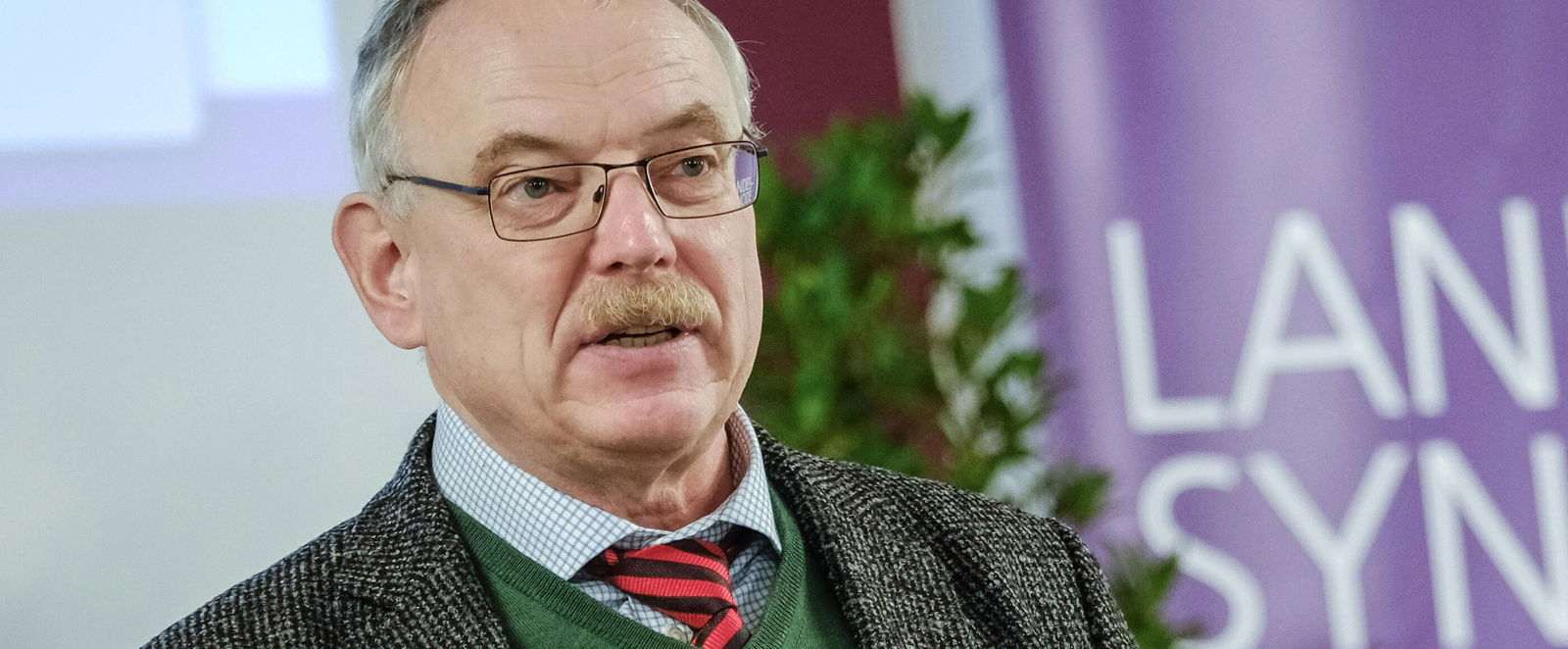 Eine männlich lesbare Person mit kurzen grauen Haaren und Brille am Rednerpult.