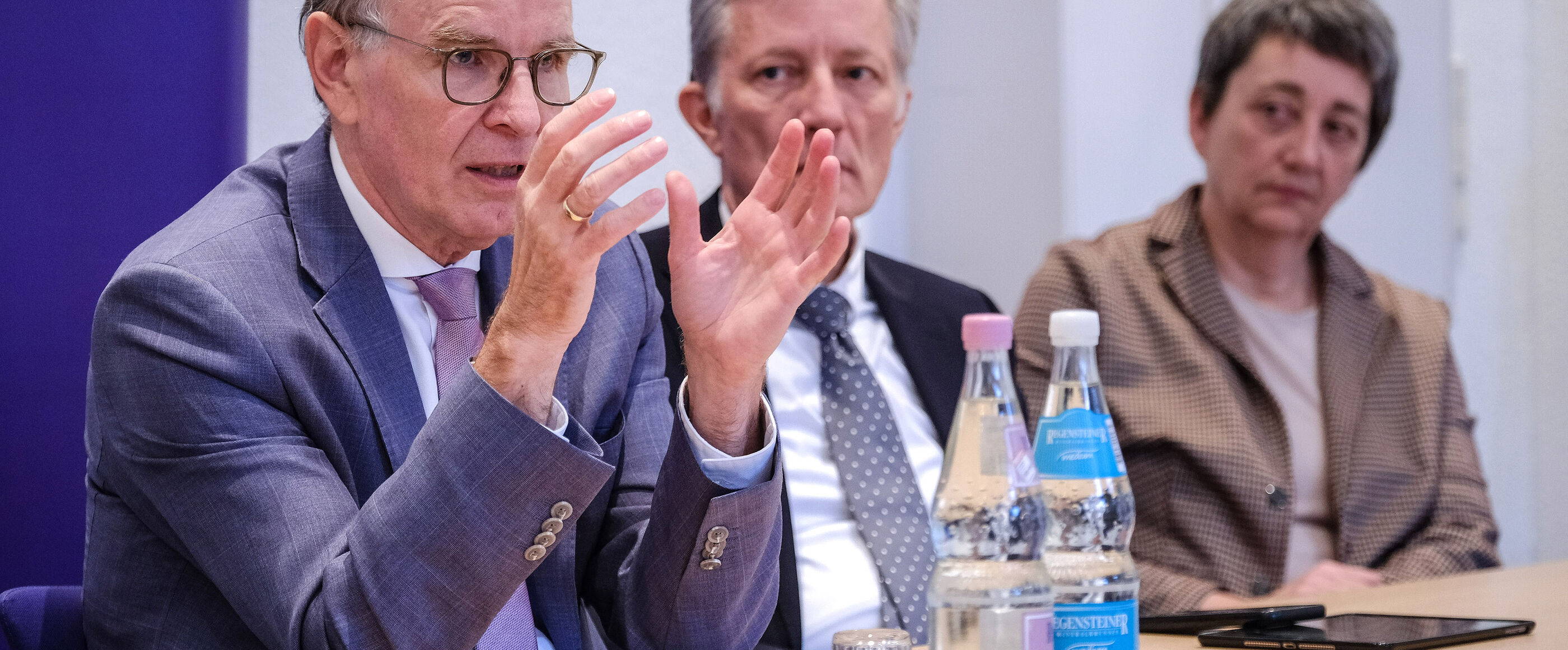 Eine männlich lesbare Person sitzt im Anzug am Tisch und gestikuliert in der Diskussion. Im Hintergrund sind weitere Personen am Tisch erkennbar.