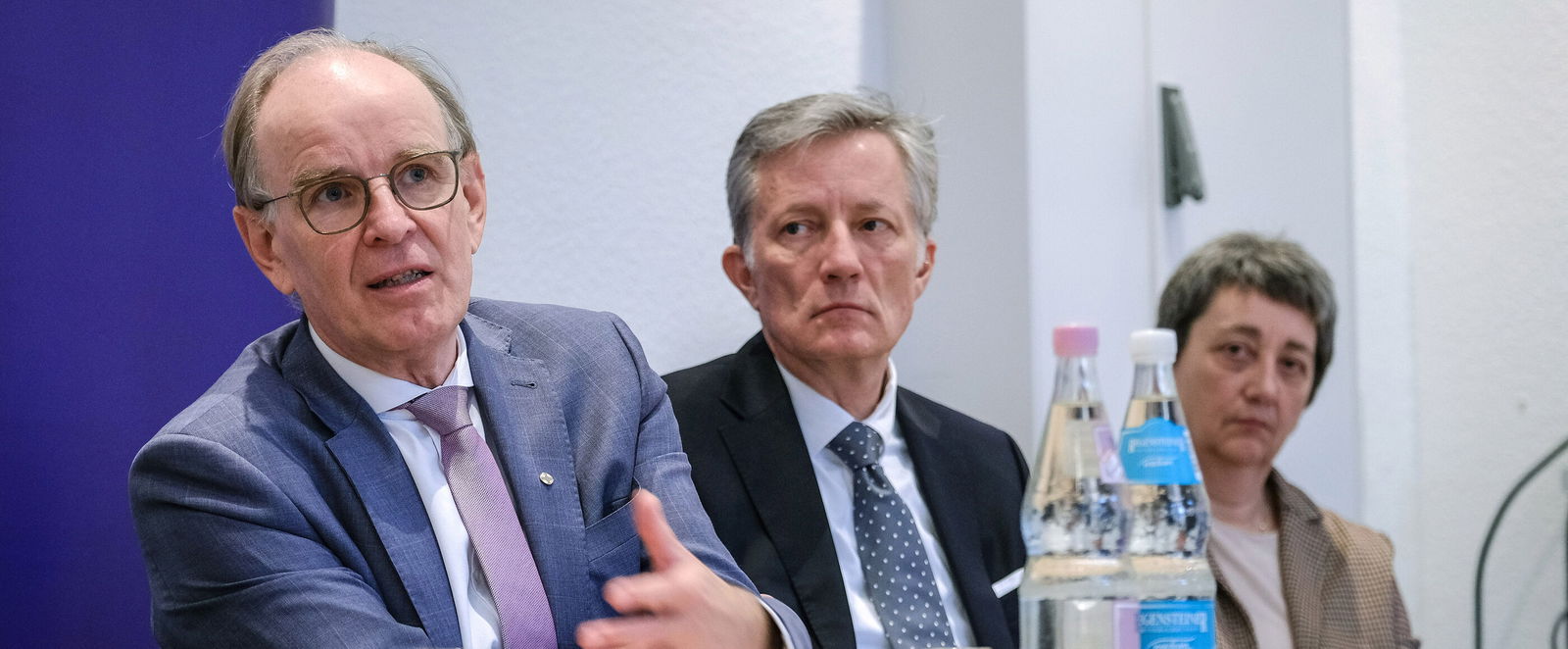 Eine männlich lesbare Person in blauem Anzug sitzt am Tisch und gestikuliert im Gespräch; im Hintergrund hören zwei weitere Personen zu.