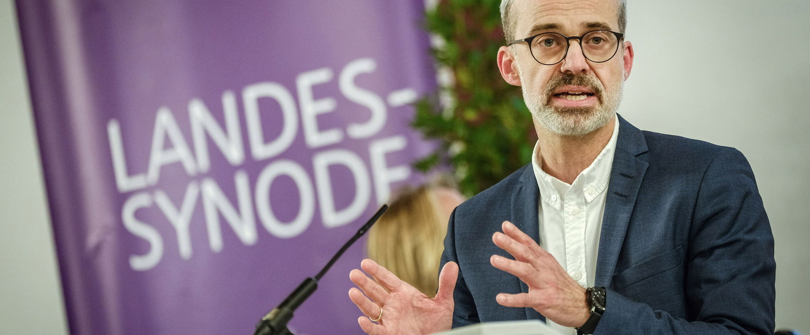 Eine männlich lesbare Person steht an einem Rednerpult und spricht.