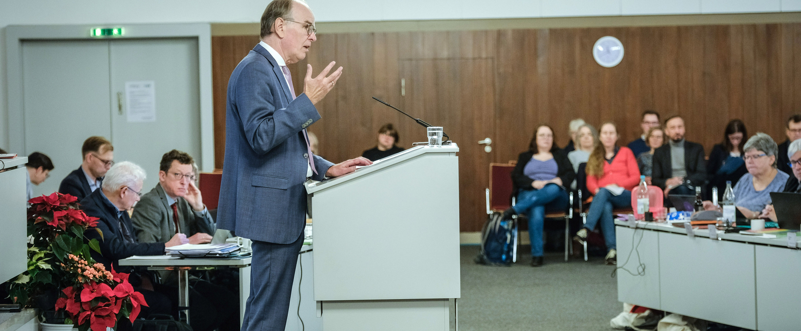 Eine männlich zu lesende Person an einem Redepult vor sitzenden Menschen.