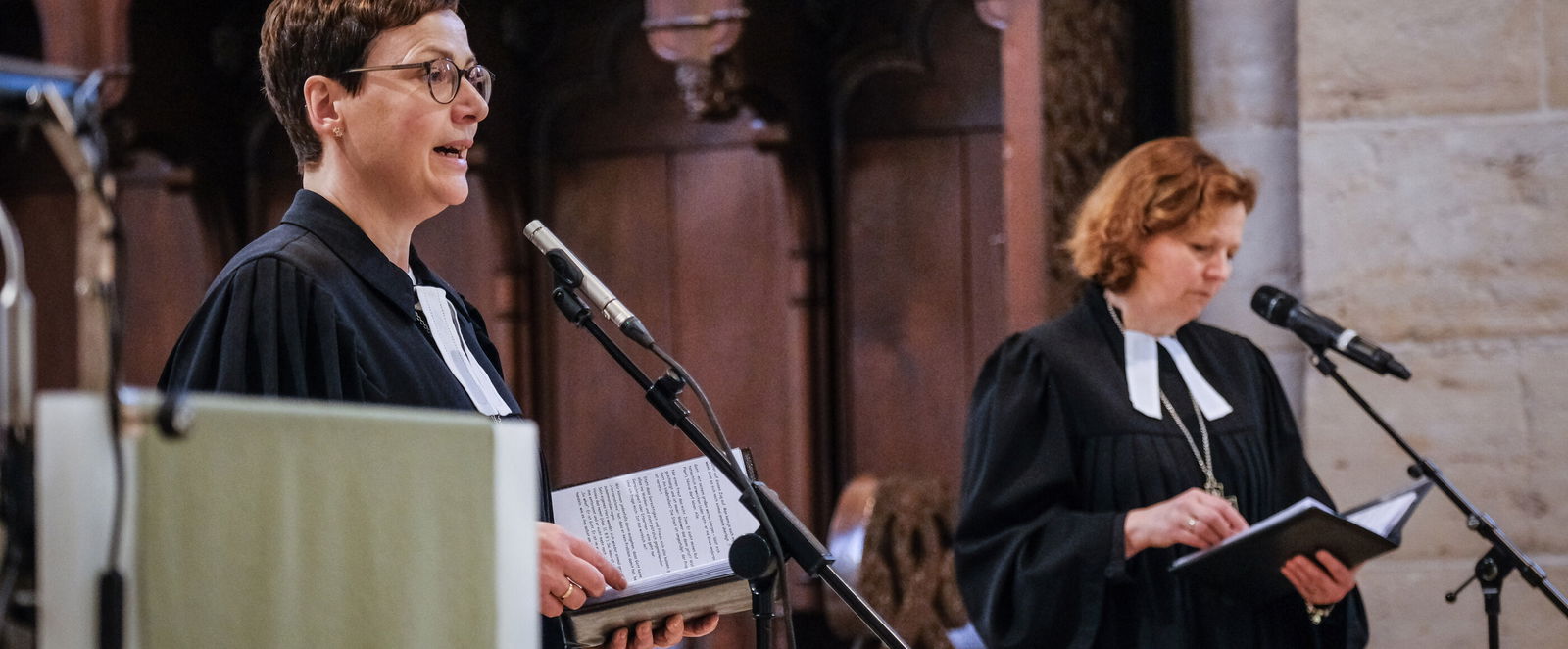 Zwei weiblich gelesene Personen mit Talar sprechen jeweils in ein Mikrofon. 