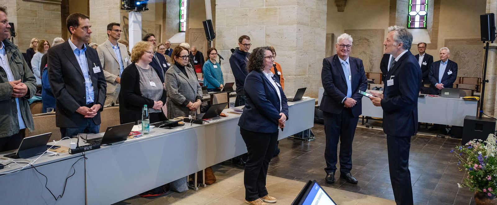 Eine Sitzung in einer Kirche, vorne stehen eine als Frau lesbare Person und zwei als Männer lesbare Personen.