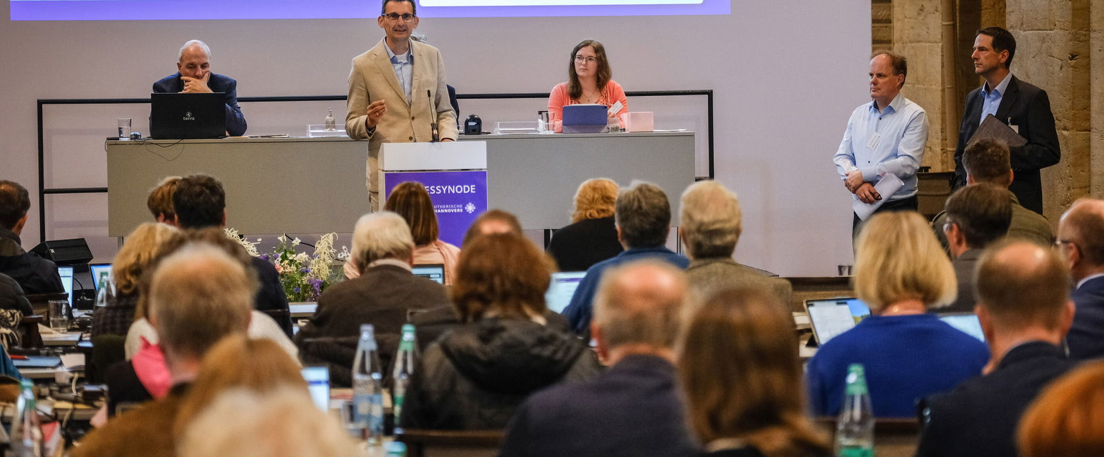 Plenumsteilnehmende sitzend im Vordergrund. Drei Personen im Fokus hinter einem Redepult.