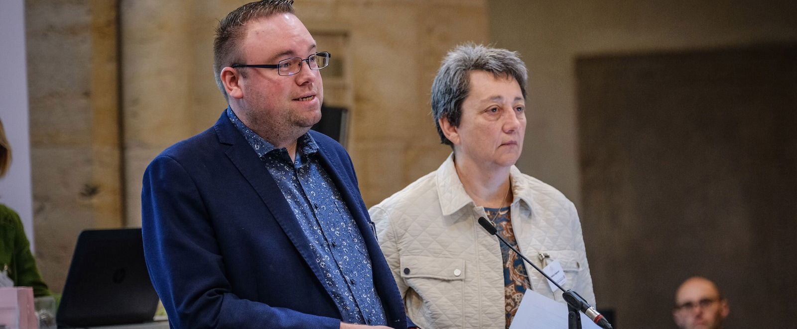 Eine weiblich und männlich lesbare Person stehen an einem Podium. Die männlich lesbare Person spricht.