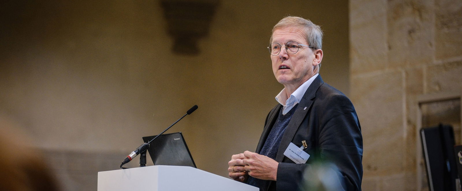 Eine männlich lesbare Person steht an einem Podium und spricht.