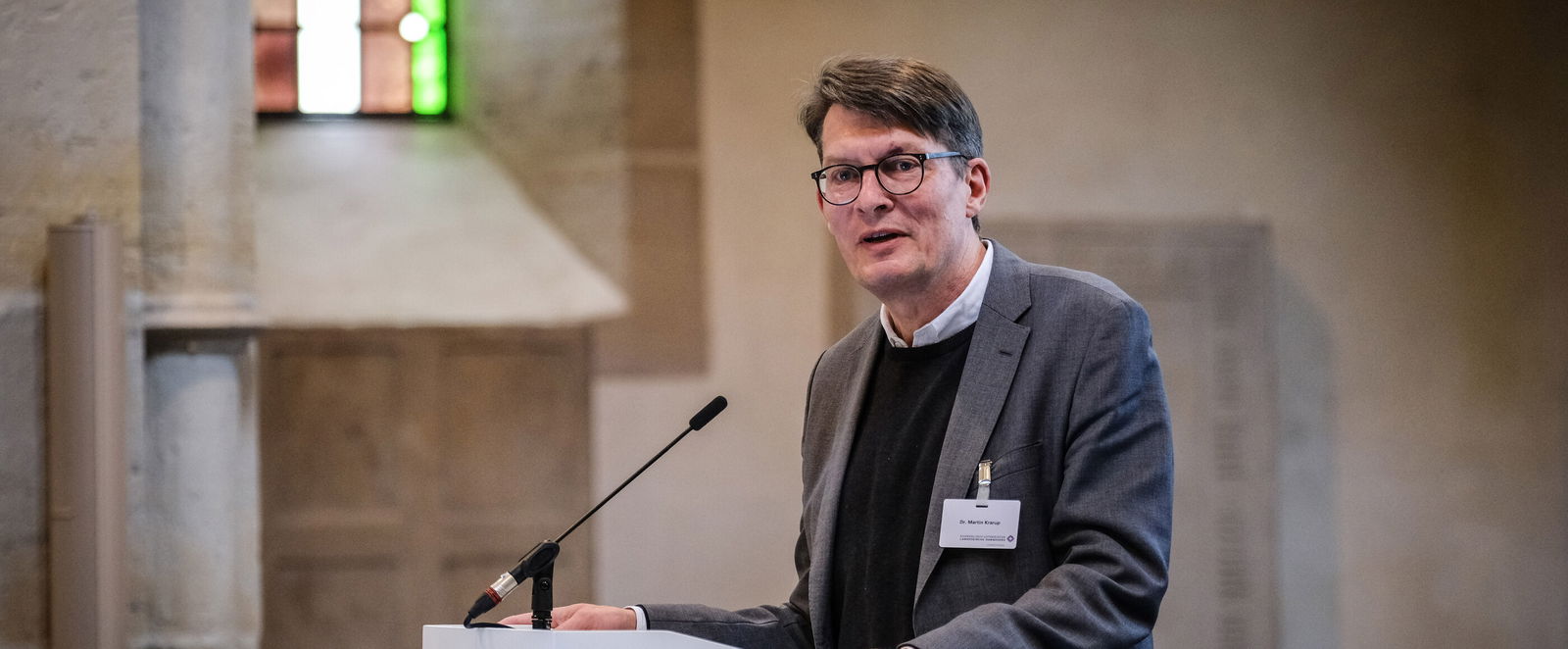 Eine männlich lesbare Person steht an einem Podium und spricht.