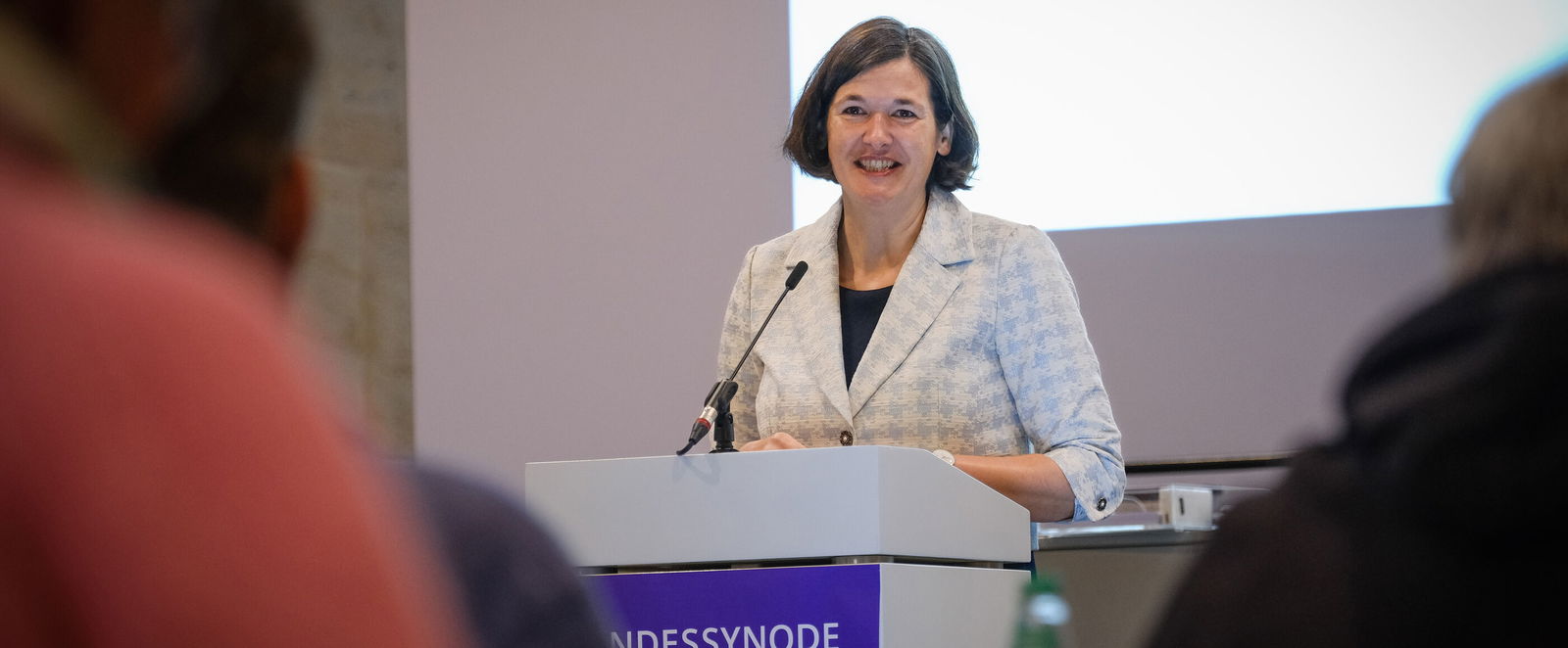 Eine als Frau lesbare Person steht an einem Redepult und spricht in ein Mikrofon.