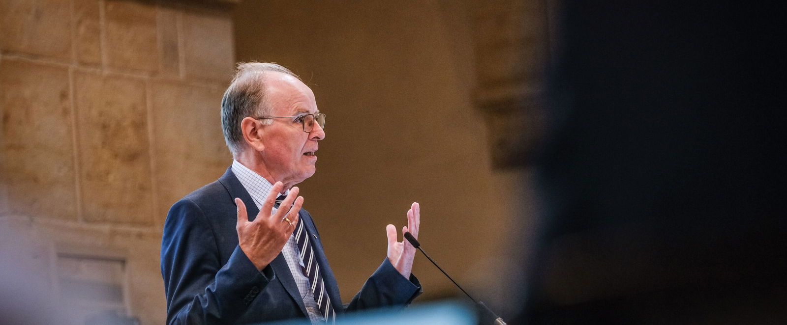 Eine männlich gelesene Person steht am Rednerpult und hebt beim Sprechen beide Hände. 