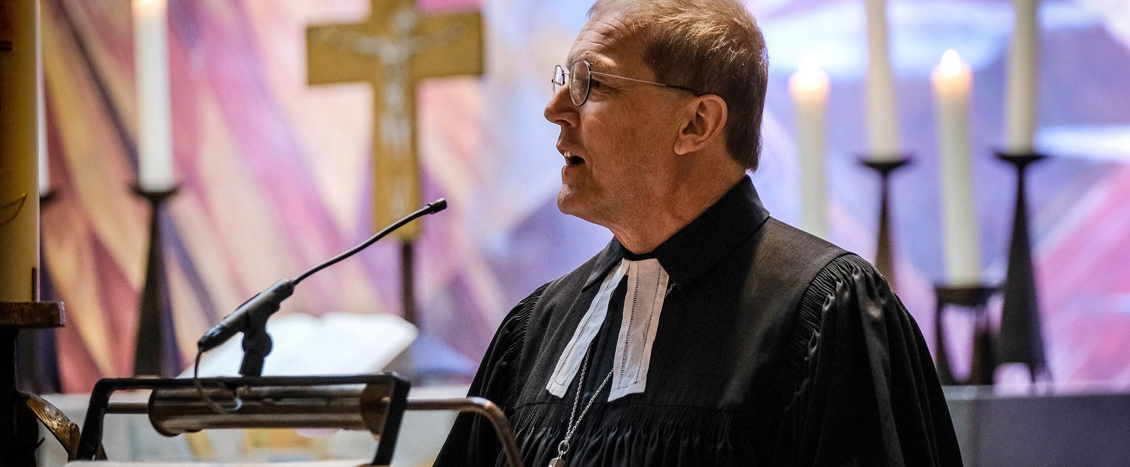 Eine als Mann lesbare Person steht in einer Kirche an einem Pult und spricht.