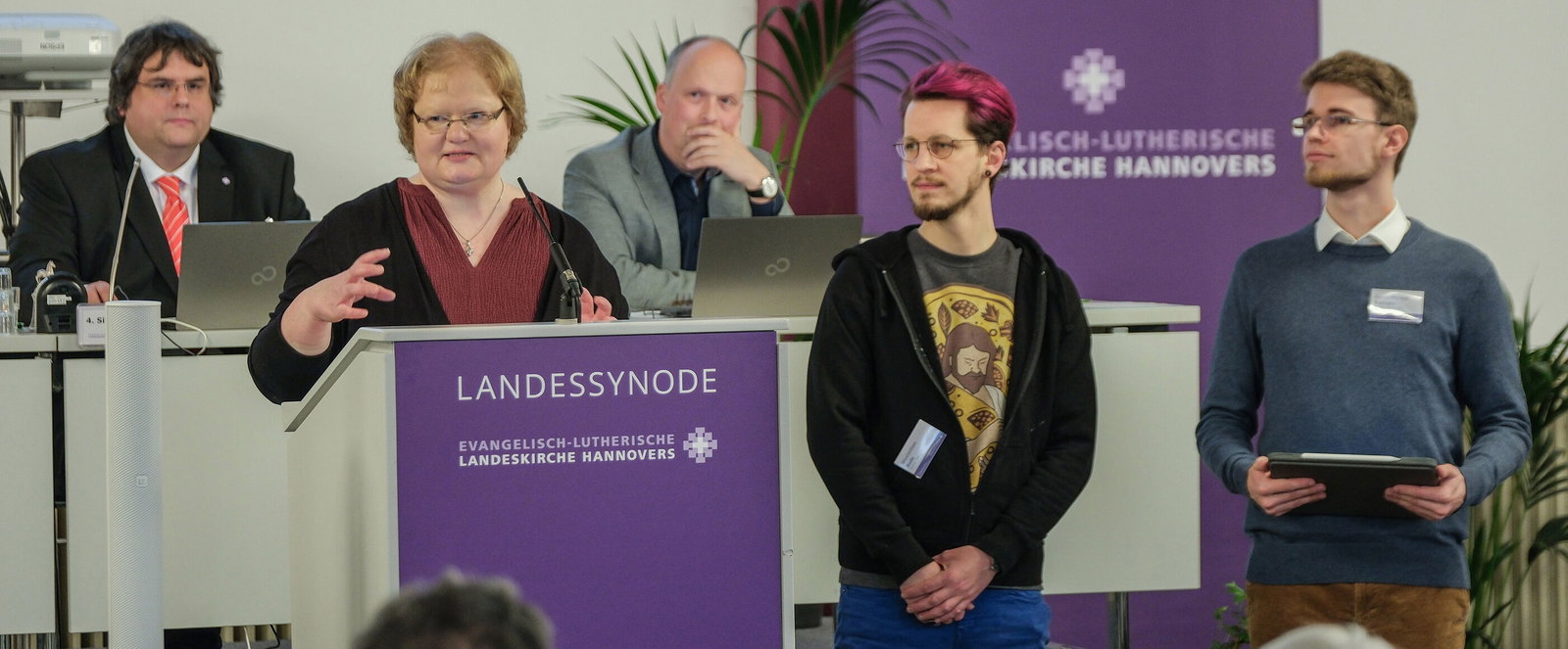 Eine Gruppe aus als Männer und Frauen lesbaren Personen an einem Redepult